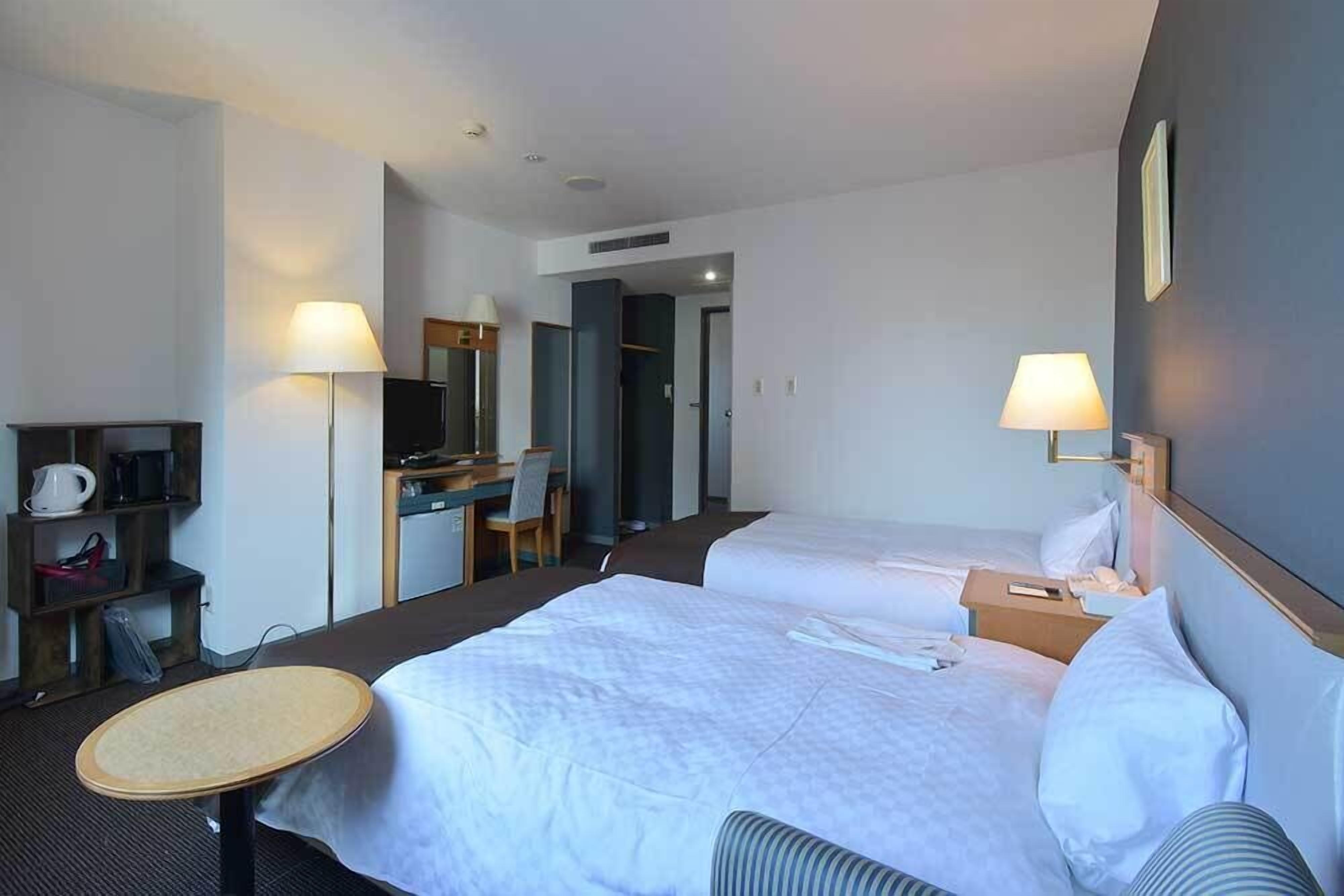 A hotel room with two beds, a desk, a chair, a lamp, and a small table.
