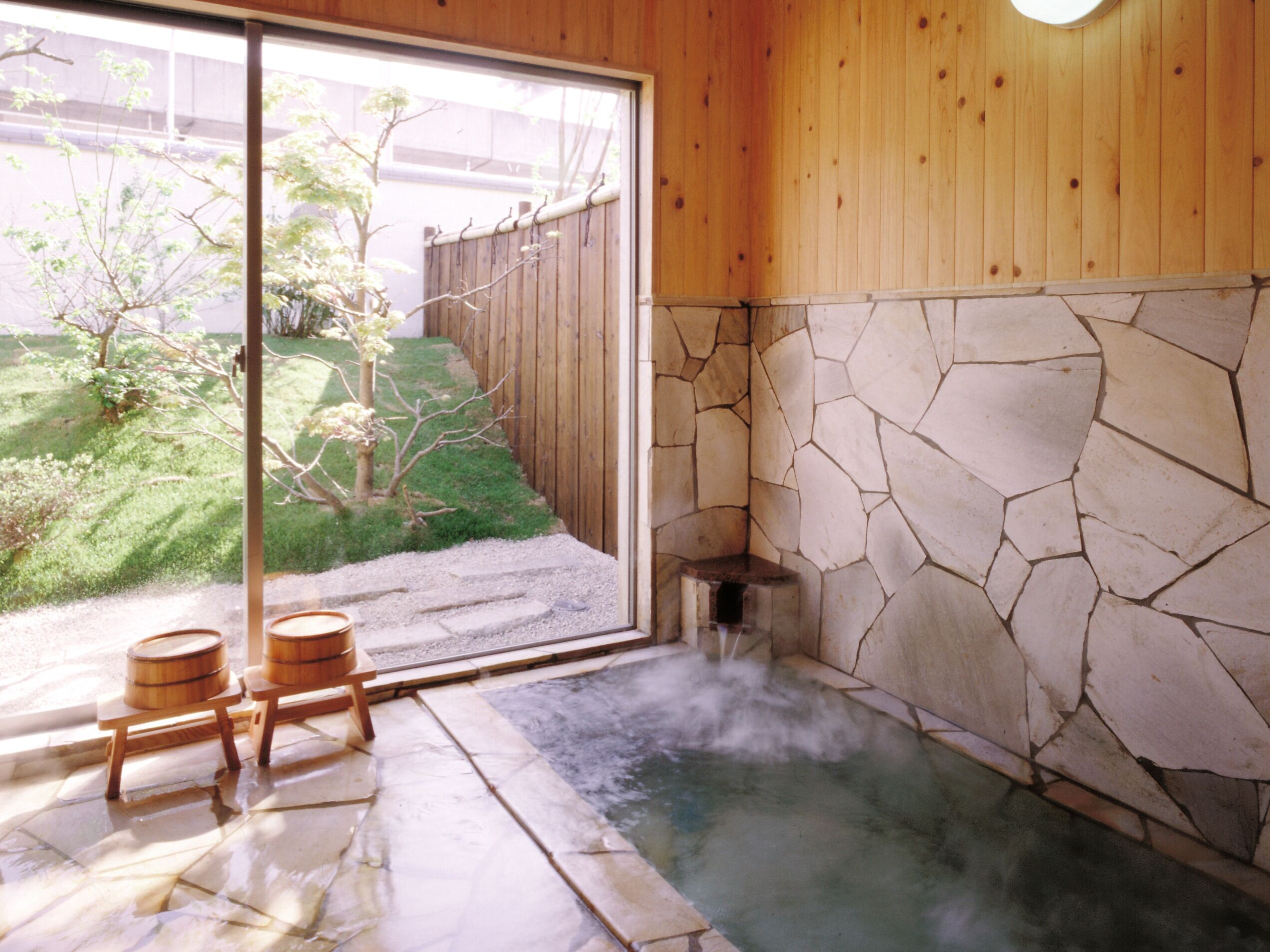 A hot spring bath with stone walls and wooden flooring, a wooden fence, and a view of a garden outside.