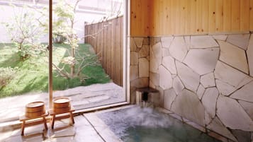 A hot spring bath with stone walls and wooden flooring, a wooden fence, and a view of a garden outside.