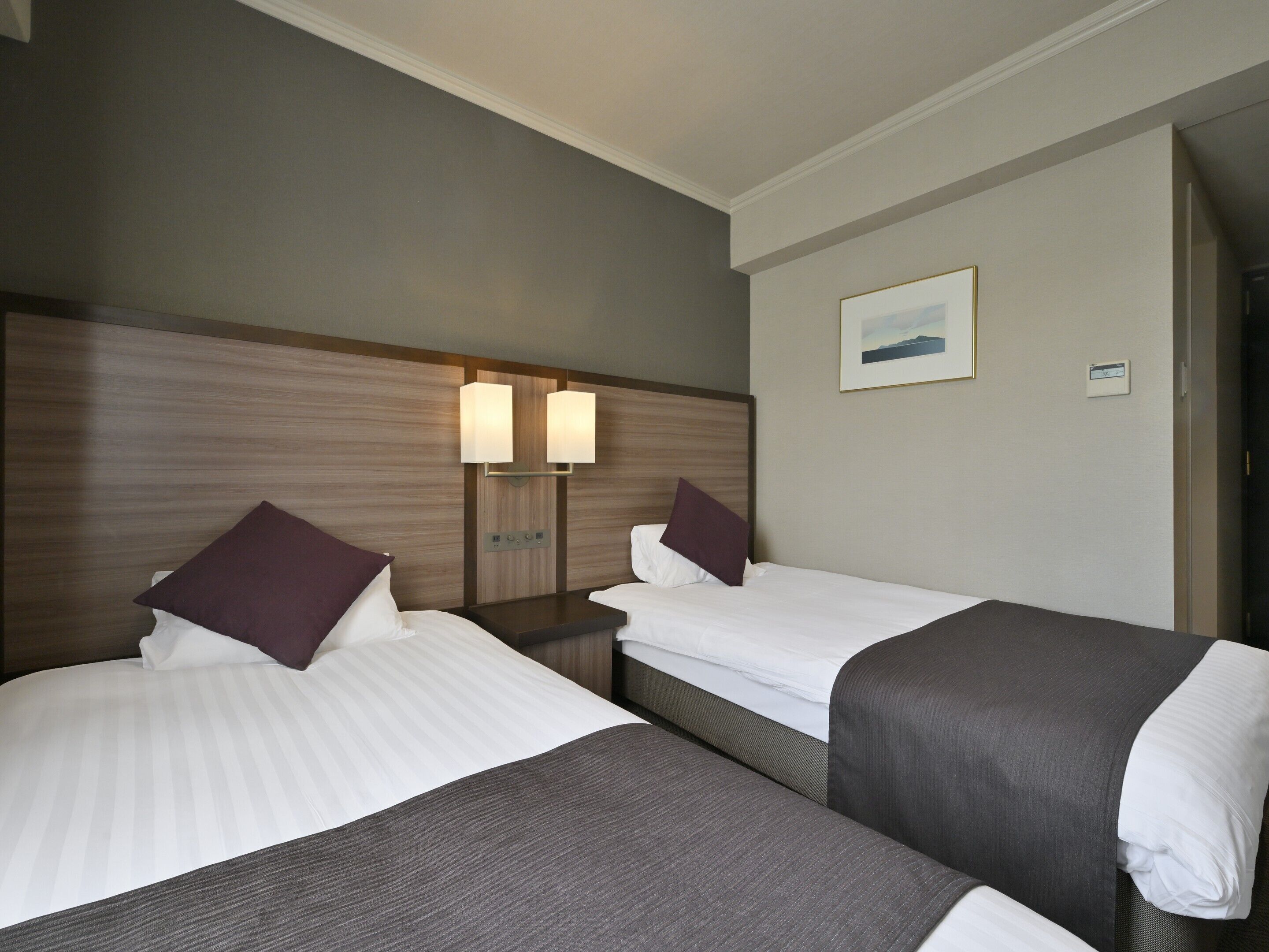 Habitación de hotel con dos camas, cabecero con luces de pared, mesita de noche pequeña y un cuadro enmarcado en la pared.