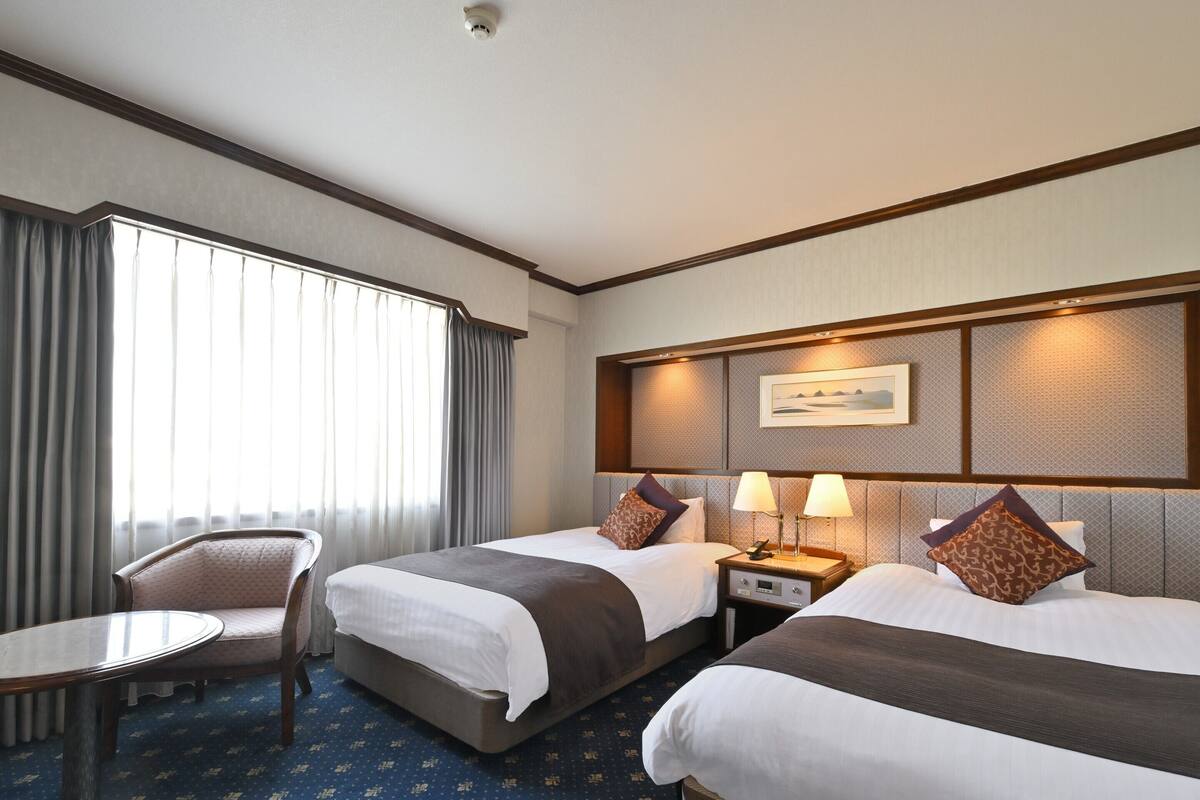 A hotel room with two beds, a small round table, a chair, and a large window with curtains.