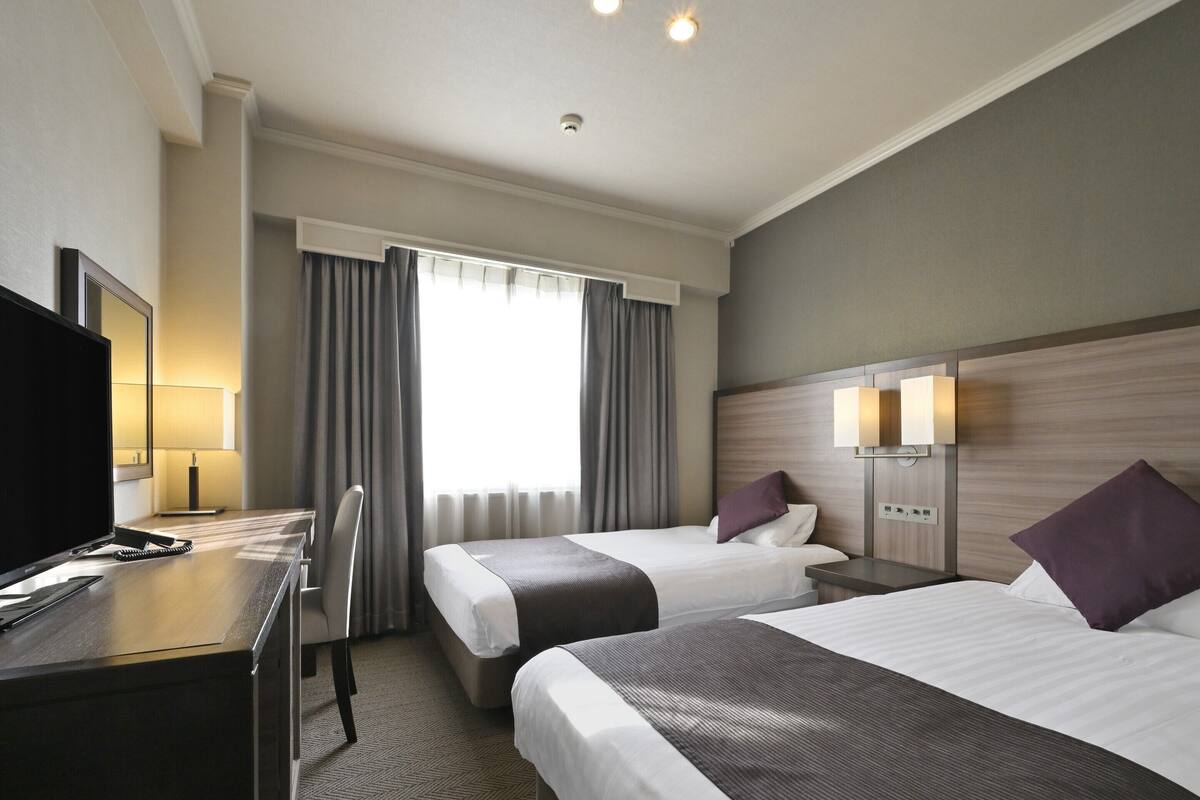 A hotel room with two beds, a desk with a computer, a TV, and a window with curtains.