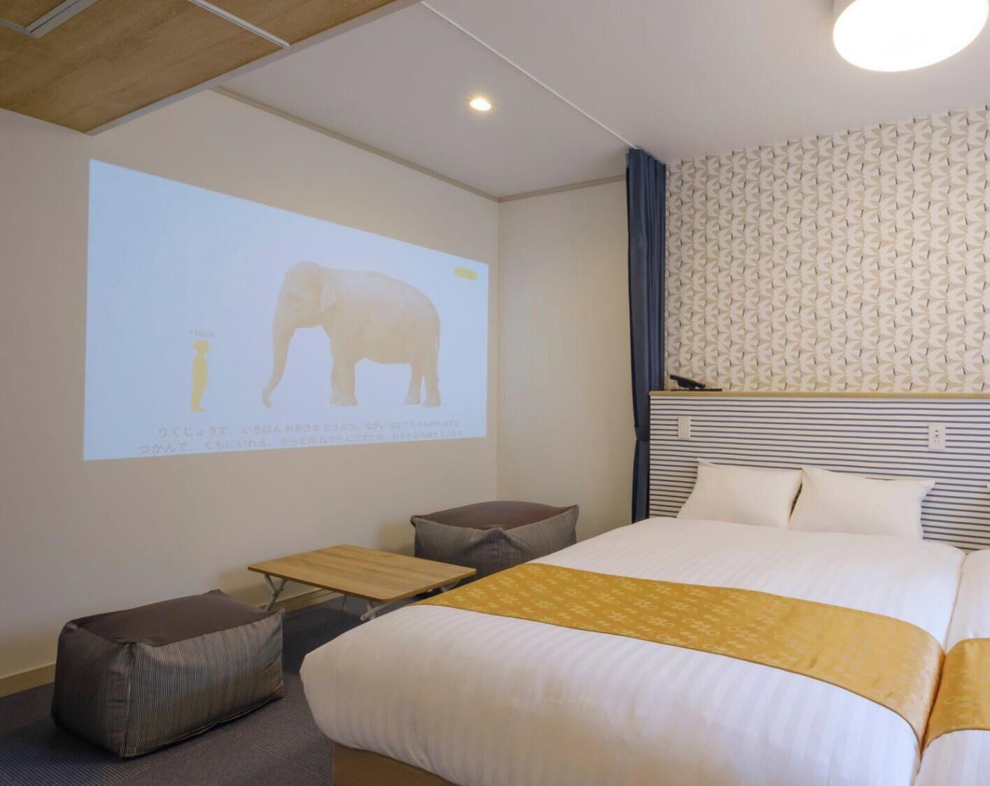 A hotel room with a bed, a small table, a projector screen displaying an elephant, and a patterned wall.