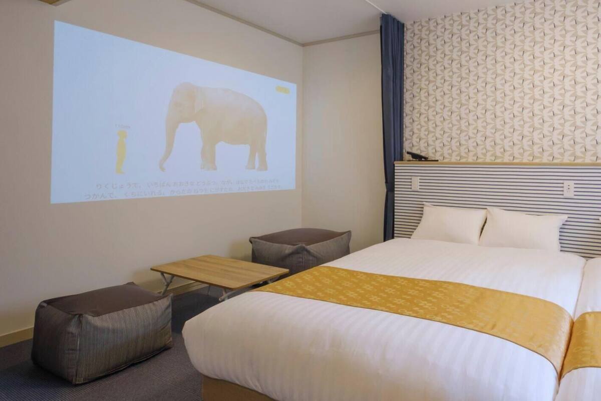 A hotel room with a bed, a small table, a projector screen displaying an elephant, and a patterned wall.
