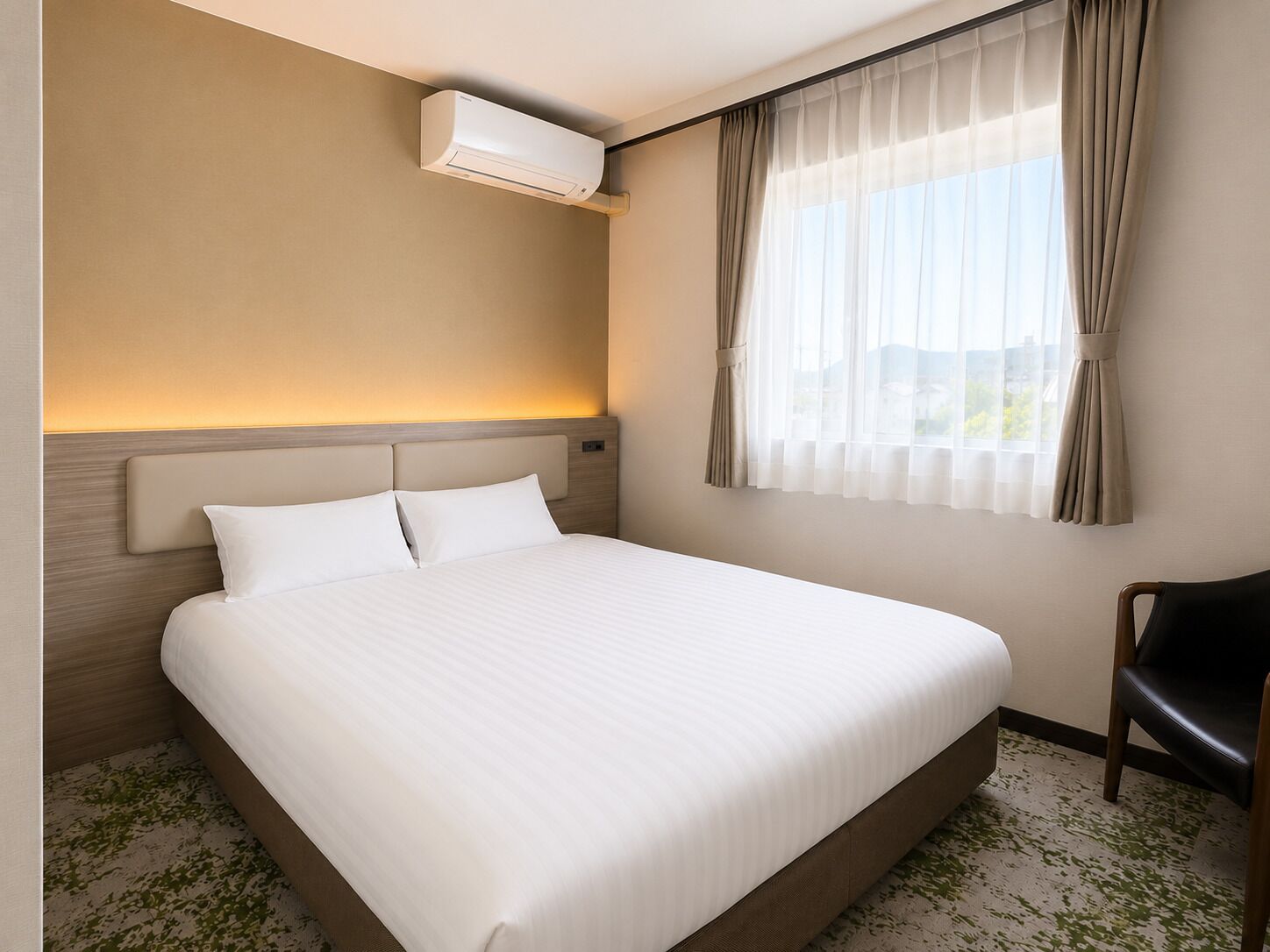 A hotel room with a large bed, an air conditioning unit, a window with sheer curtains, and a view of the outdoors.