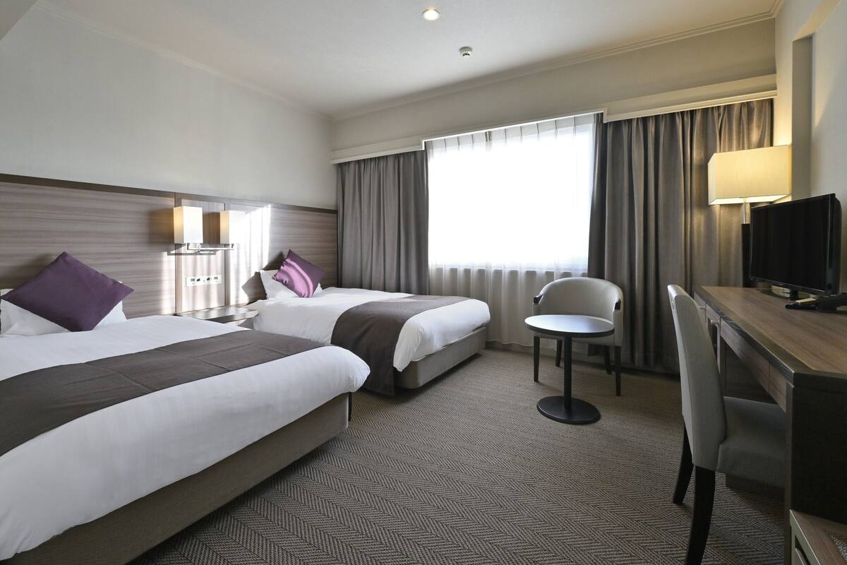 A hotel room with two beds, a desk with a chair, a television, and a window with curtains.