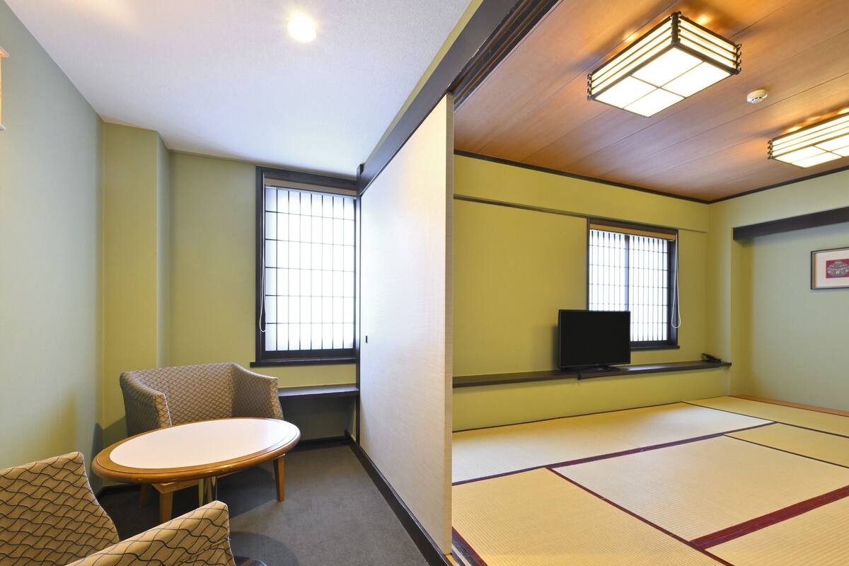 A room with tatami mats, a tatami mat table, two tatami mat chairs, a television on a shelf, and two windows with blinds.