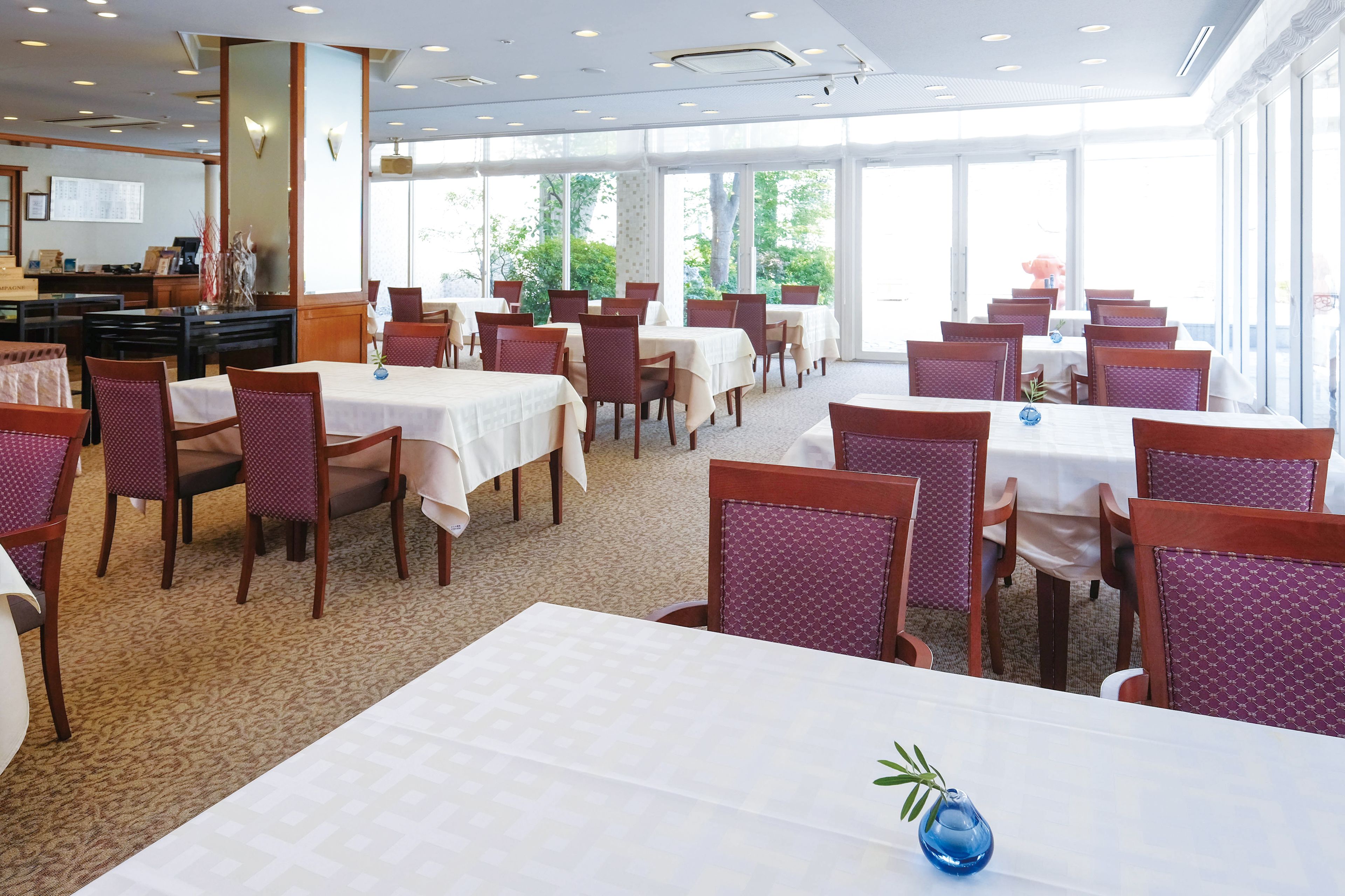 Un espace repas avec des tables et des chaises, de grandes fenêtres et un sol recouvert de moquette.