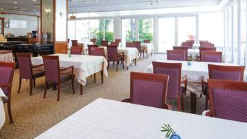 Un espace repas avec des tables et des chaises, de grandes fenêtres et un sol recouvert de moquette.