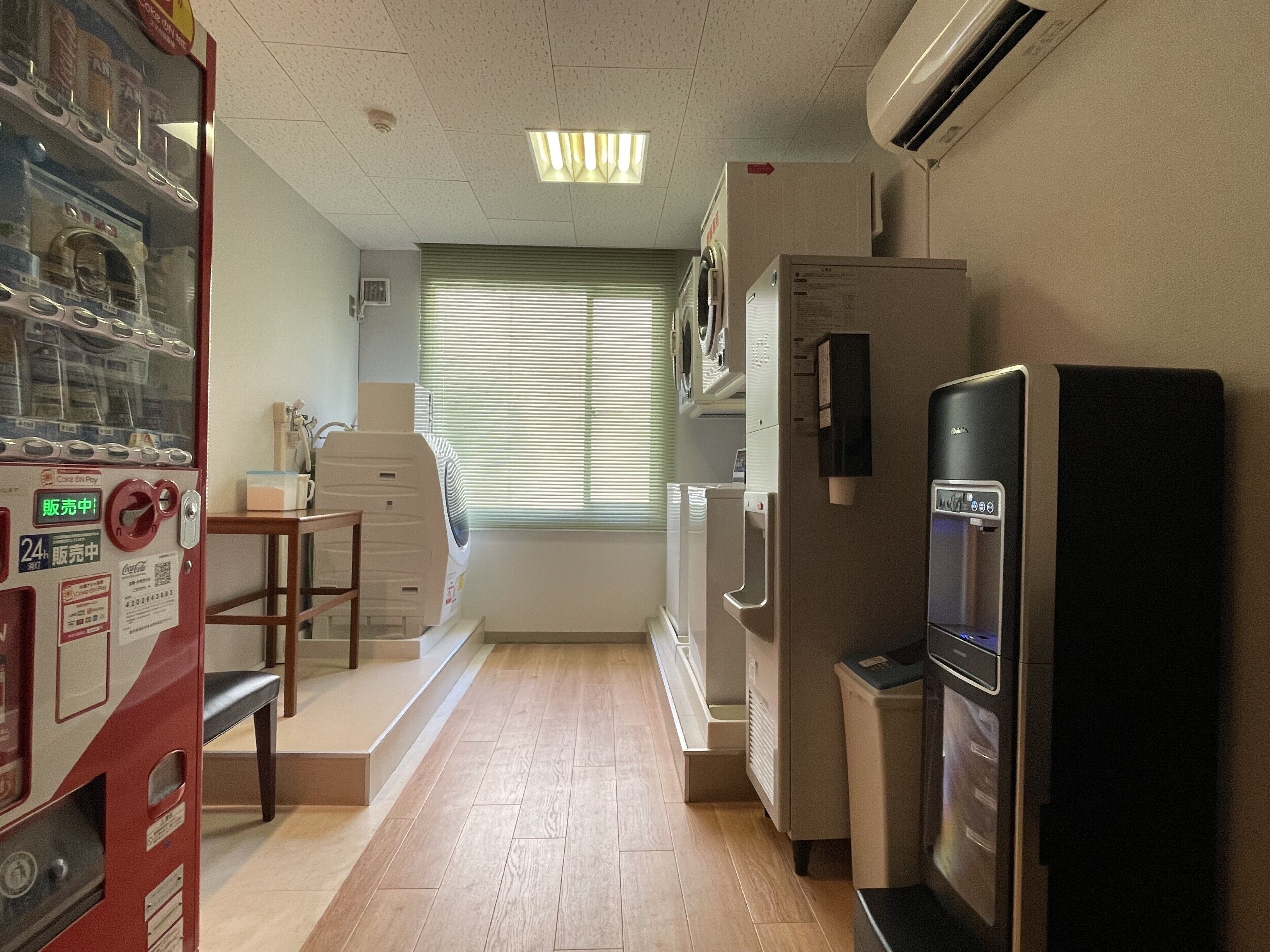 A compact hotel room with a kitchenette, a small table, a chair, a window with blinds, and a refrigerator.