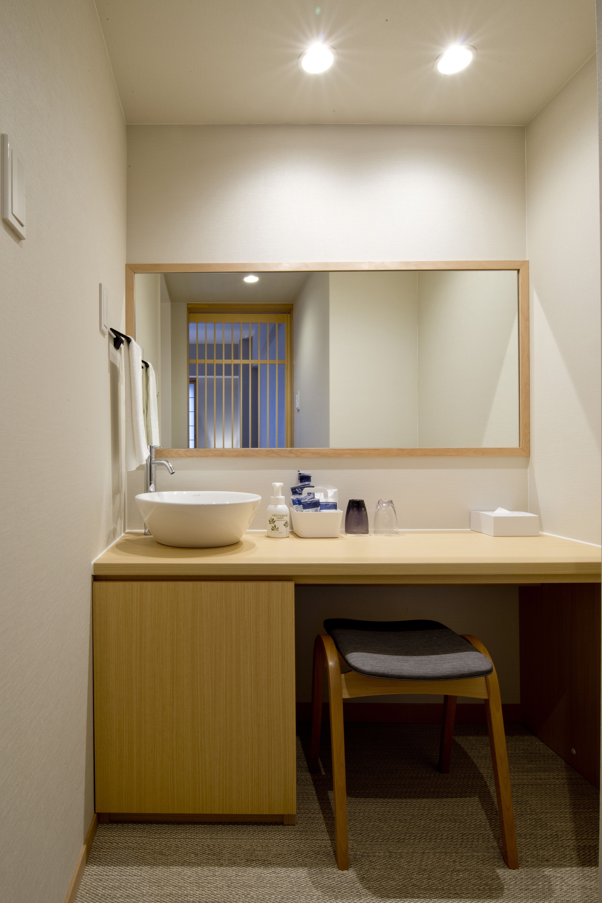 Un baño con lavamanos blanco, un espejo, un perchero para toallas y una silla de madera.