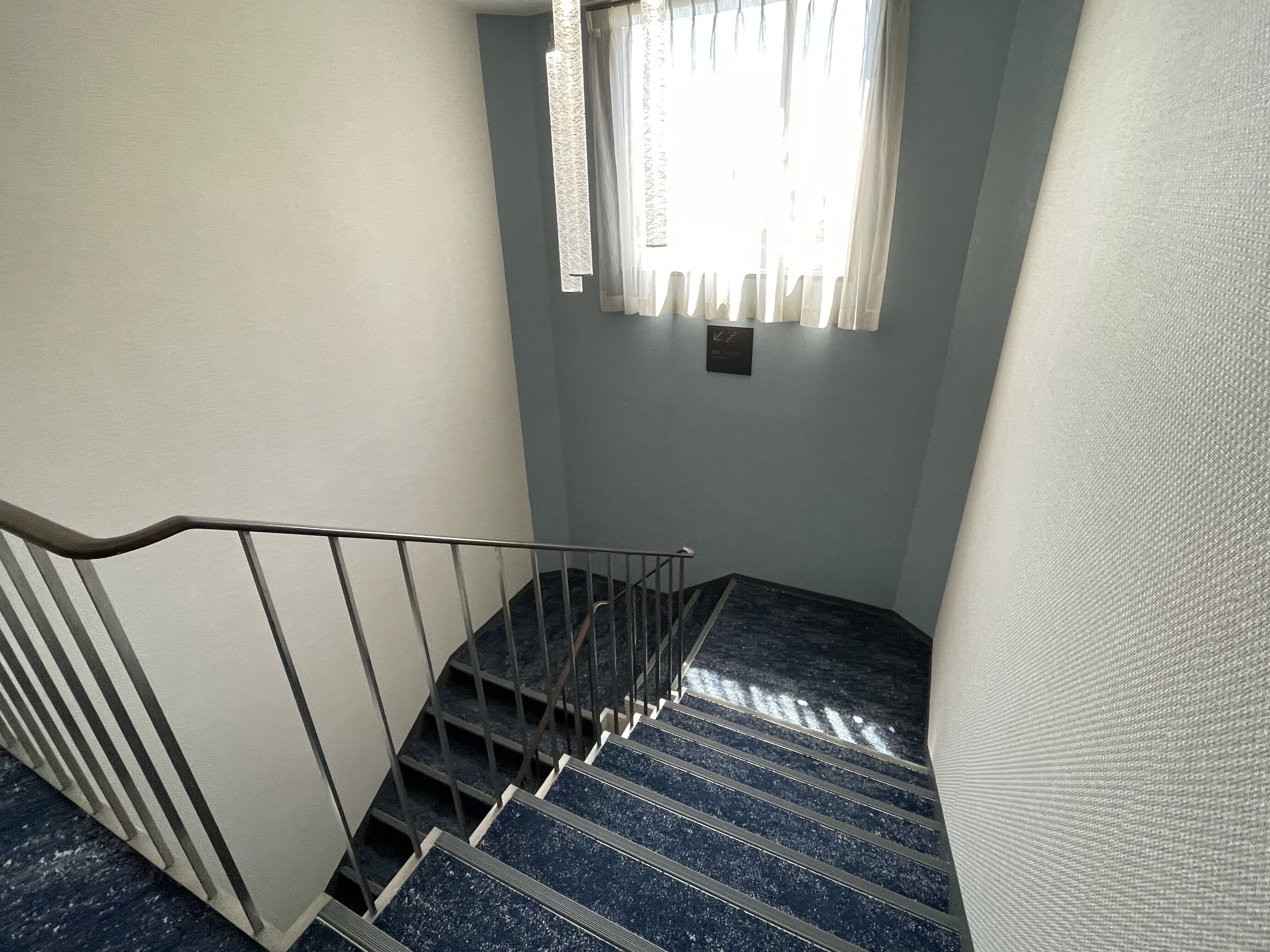 Una escalera con barandales de metal y pasamanos, que lleva a un rellano con un interruptor de luz y un reloj en la pared.