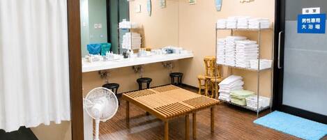 A hotel room with a wooden floor, a fan, a table, a mirror, a clock, and a towel rack.
