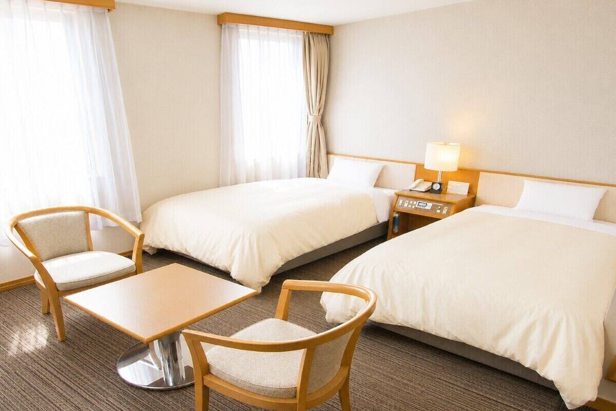 A hotel room with two beds, a small table, two chairs, a nightstand with a lamp, and a window with curtains.