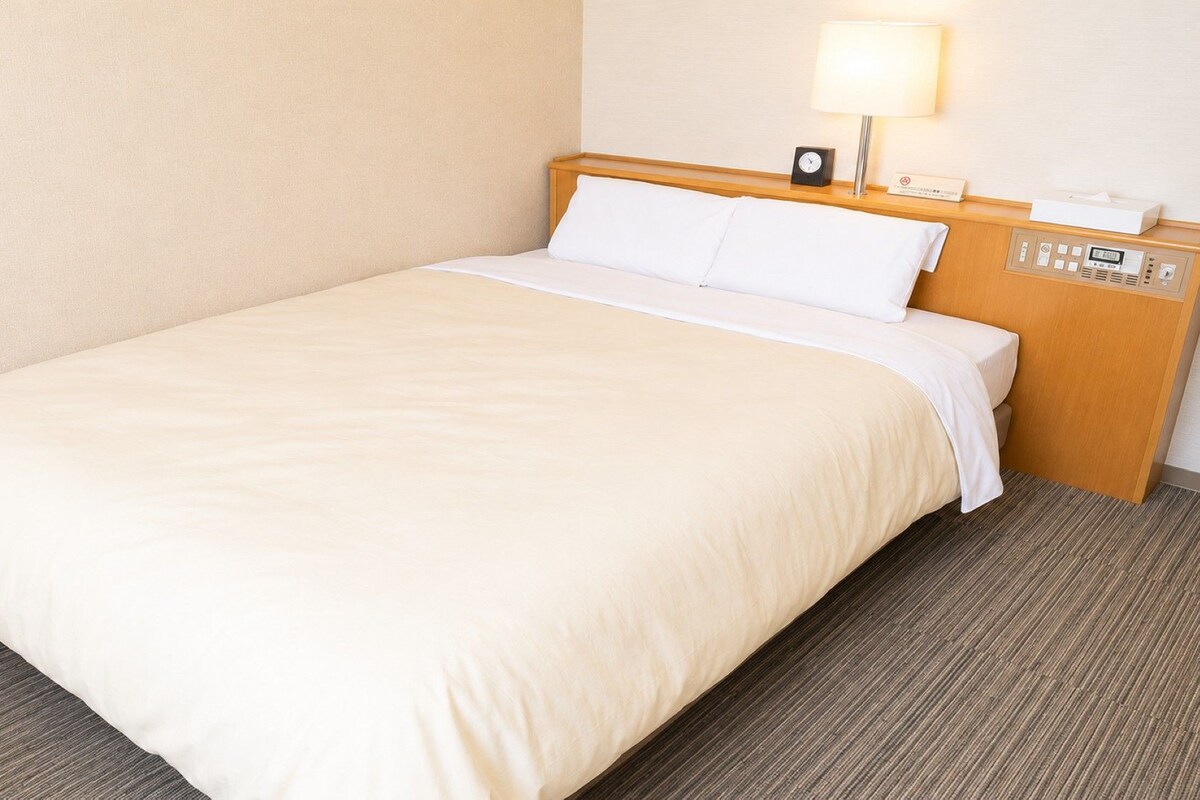 A hotel room with a bed, a nightstand with a lamp, and a control panel.