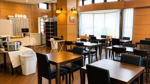 Een restaurantruimte in een hotel, met houten tafels en stoelen, een keuken met apparatuur, en grote ramen voorzien van gordijnen.