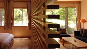 Ein modernes Schlafzimmer mit einem Bett, einem Schlafsofa und einem Couchtisch. Große Fenster mit Vorhängen bieten Blick auf viel Grün.