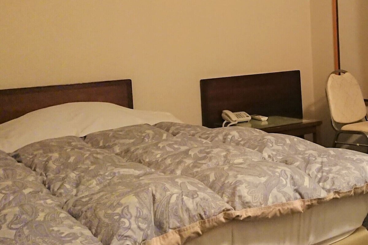 A hotel room with a bed, a bedside table with a phone, and an air conditioning unit on the wall.