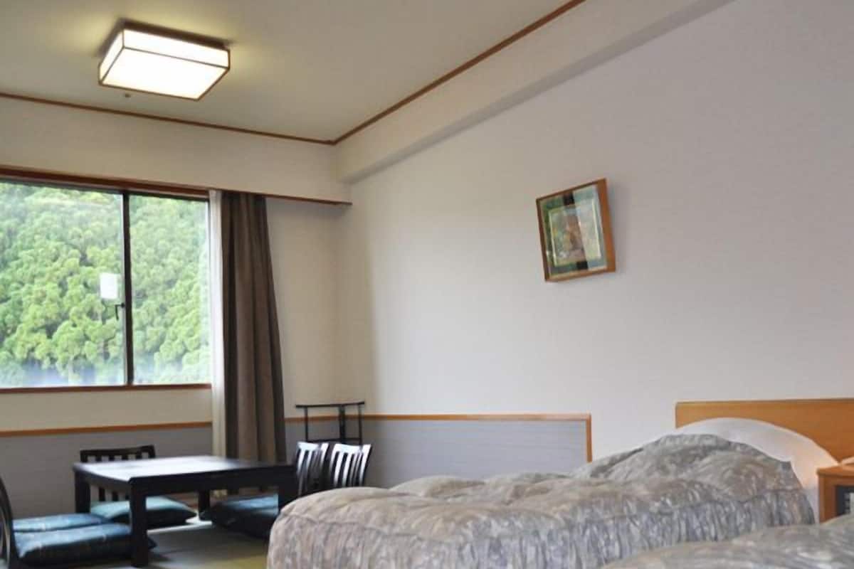 Ein Zimmer mit zwei Betten, einem Tisch, Stühlen, einem Fenster mit Blick auf Bäume und einer Deckenleuchte.