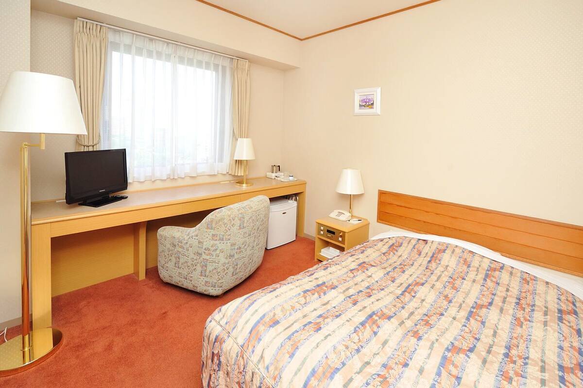 Habitación de hotel con una cama, un escritorio con computadora, una silla, una lámpara de escritorio, una mesita de noche con lámpara y un cuadro pequeño en la pared.