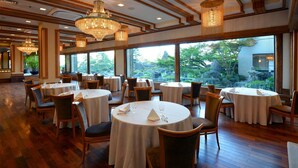 Een restaurant met ronde tafels, stoelen en kroonluchters. Grote ramen bieden uitzicht op groen buiten.
