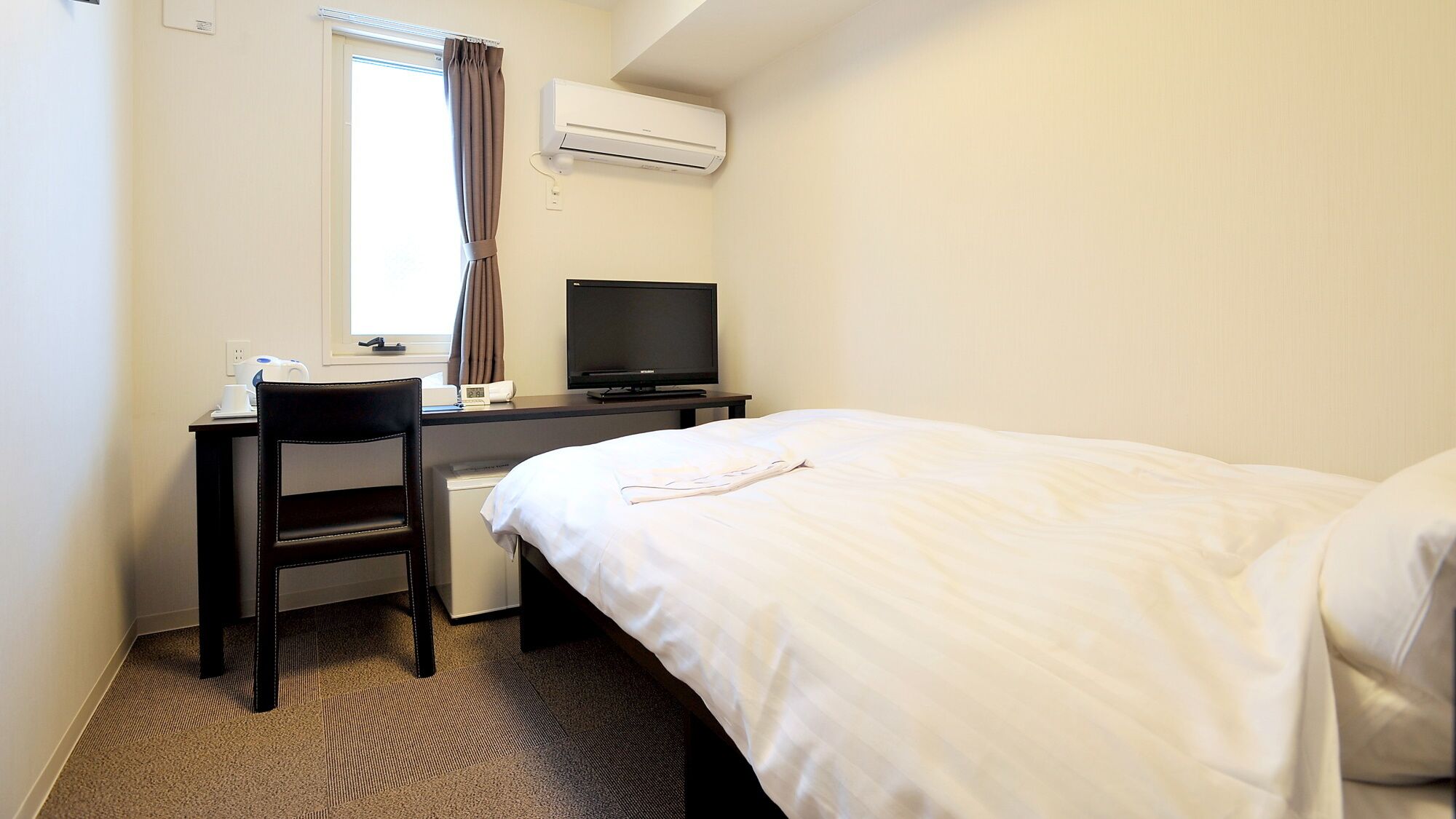 A hotel room with a bed, a desk with a chair, a television, and an air conditioning unit.