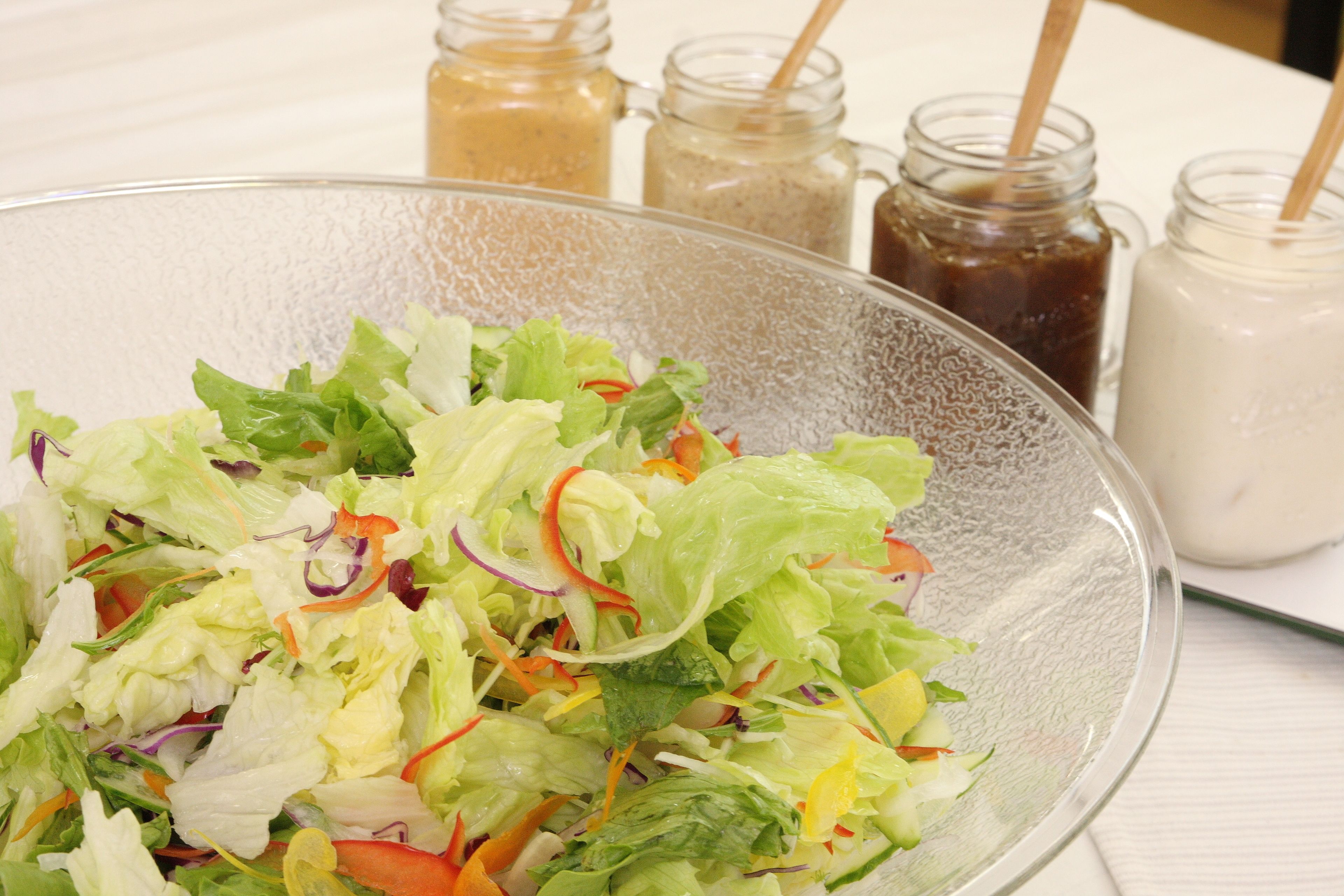 Een glazen schaal met een gemengde salade en diverse groenten, op een witte tafelkleed.