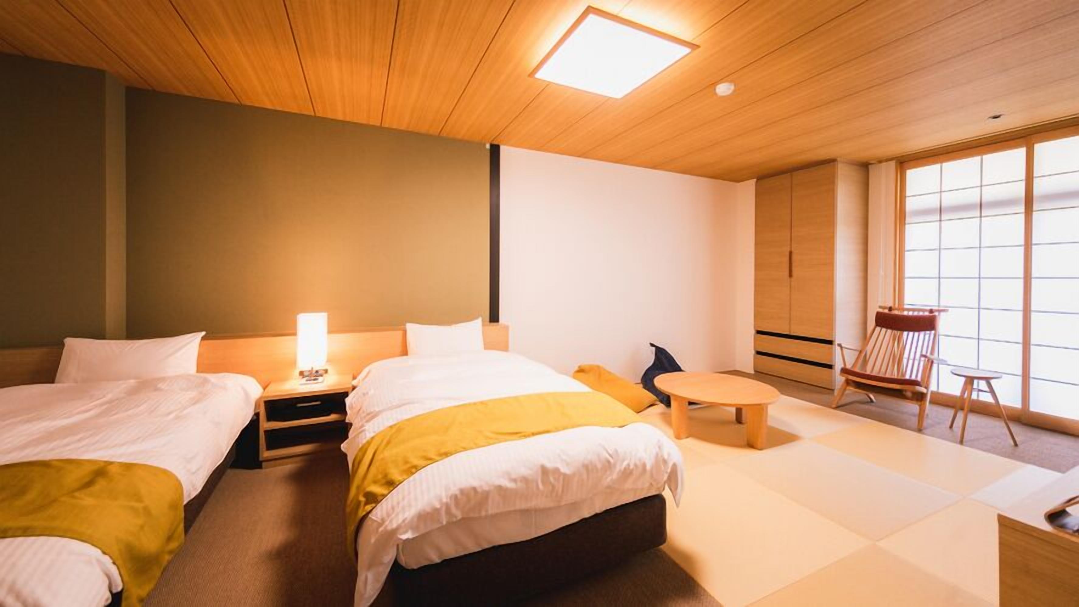 Ein Hotelzimmer mit zwei Betten, einem kleinen Tisch, einem Sessel und einem großen Fenster.