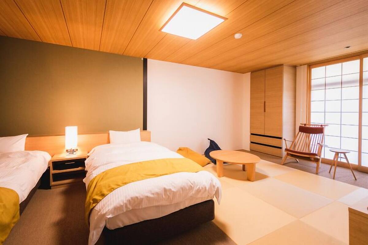 A hotel room with two beds, a small table, a chair, and a large window.