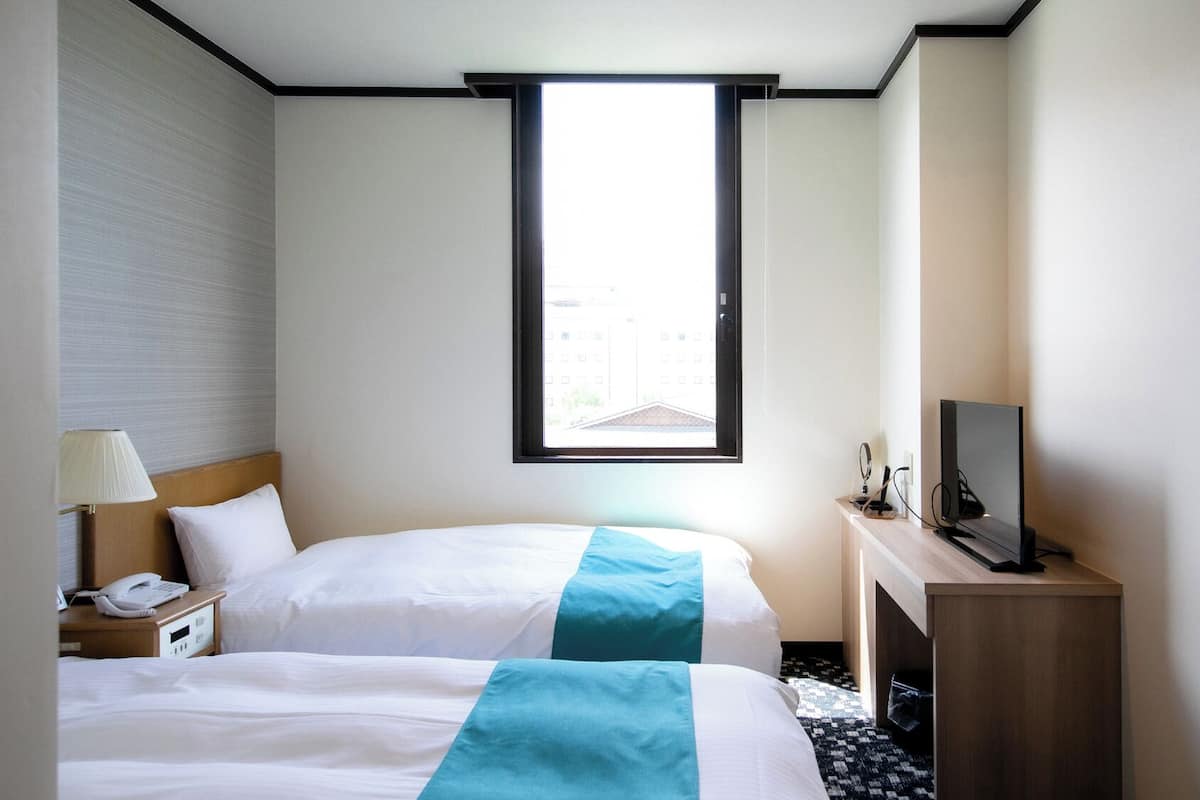 A hotel room with a bed, a desk with a TV, a phone, and a window with a view of buildings.