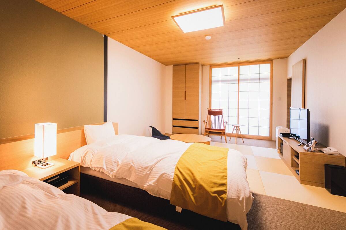 A hotel room with two beds, a desk, a chair, a TV, and a window.