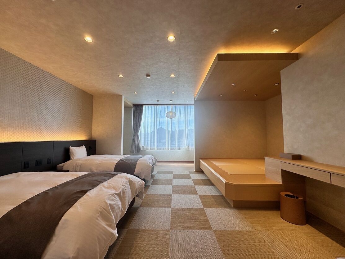 Ein modernes Hotelzimmer mit zwei Betten, einer Bank, einem Schreibtisch und einem großen Fenster mit Vorhängen.