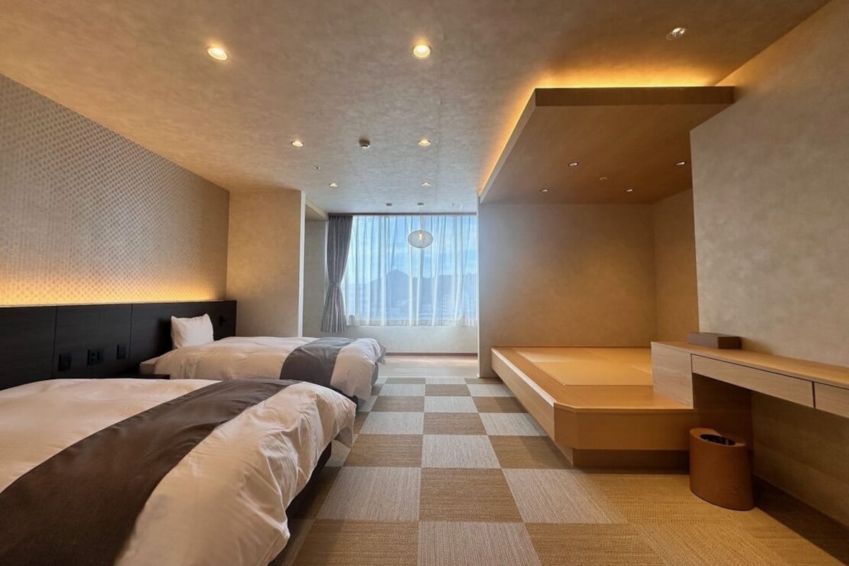 A modern hotel room with two beds, a bench, a desk, and a large window with curtains.