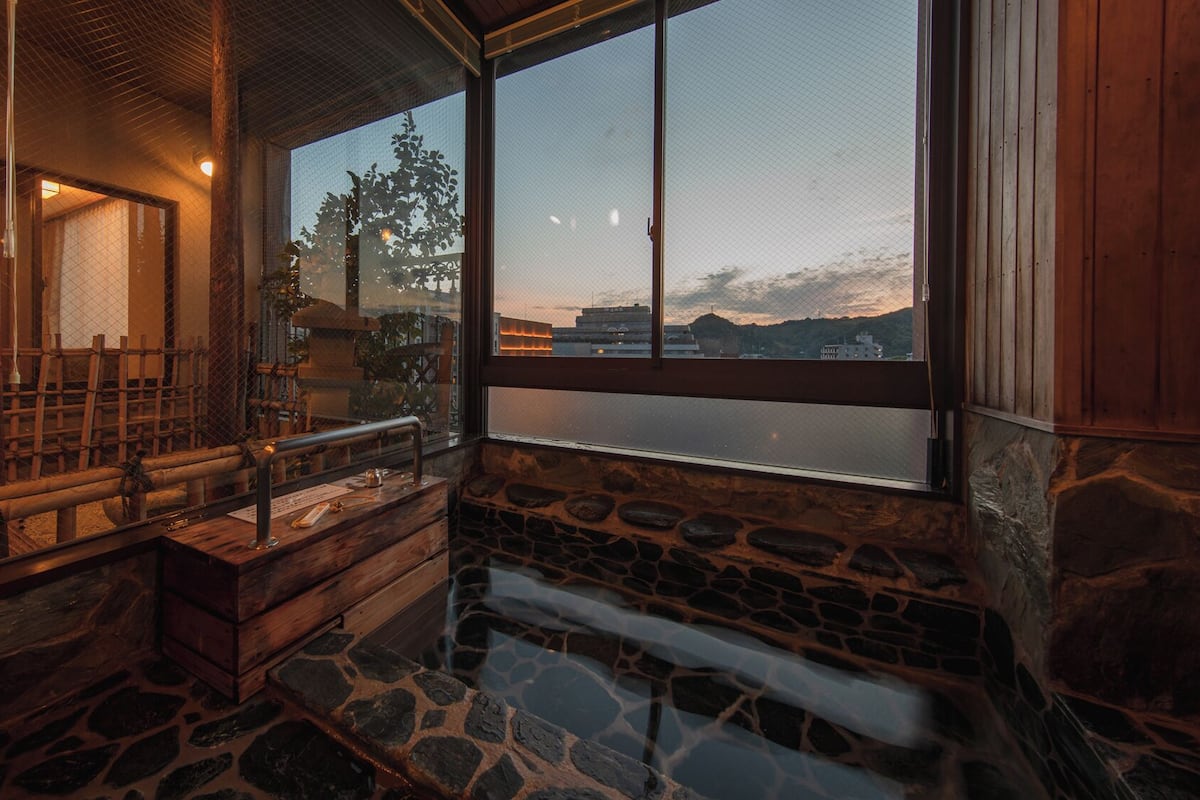 A stone-floored room with a wooden bench, a small table, and a large window offering a view of a mountain and buildings.