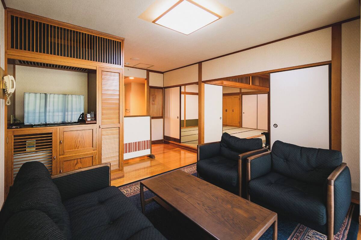 A traditional Japanese-style room with tatami flooring, a low wooden table, dark upholstered chairs, and a television.