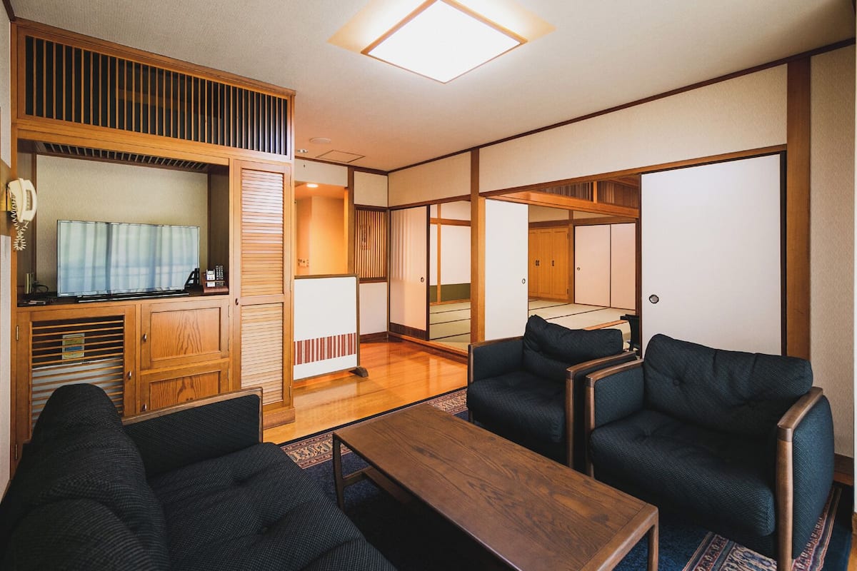 A traditional Japanese-style room with tatami flooring, a low wooden table, dark upholstered chairs, and a television.