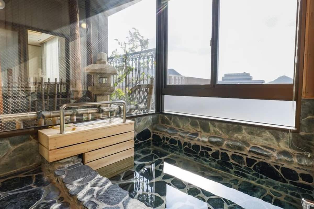 A stone-walled bathroom with a wooden bench, a sink with a metal faucet, and a view of buildings outside.