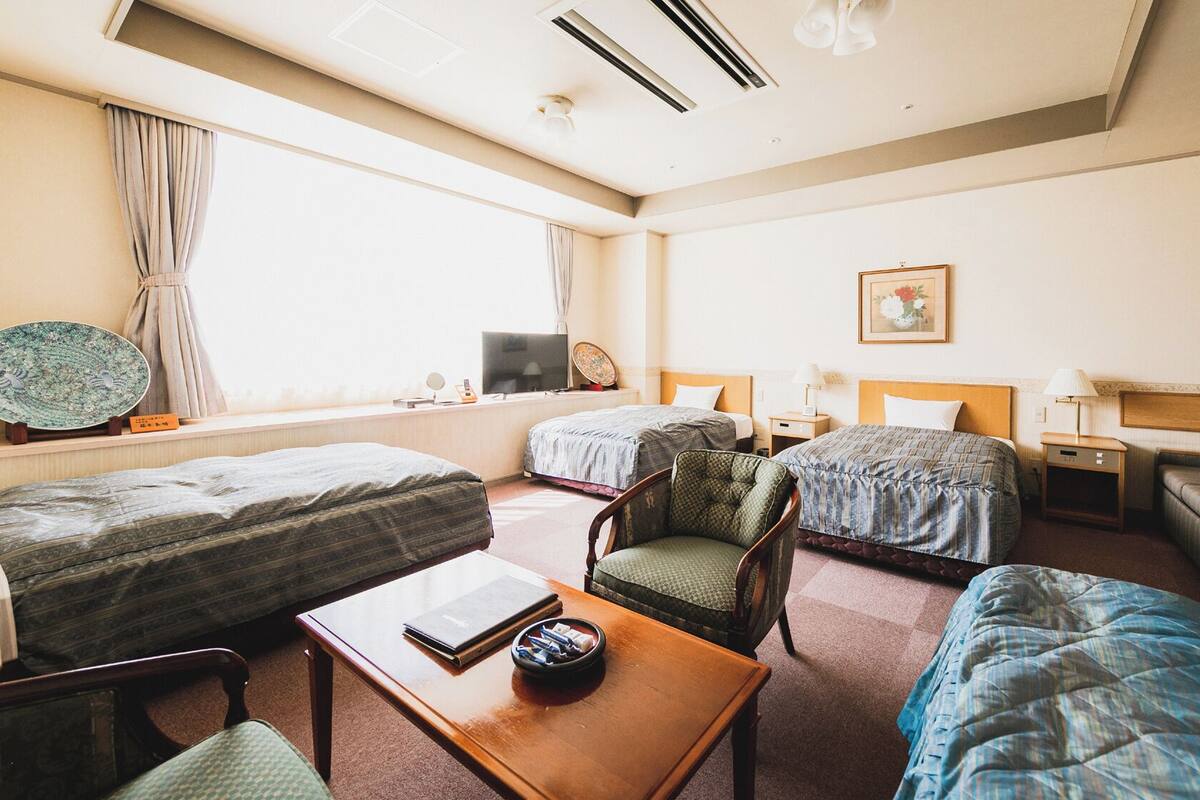 A hotel room with two single beds, a sofa, a chair, a small table, a TV, and a window with curtains.
