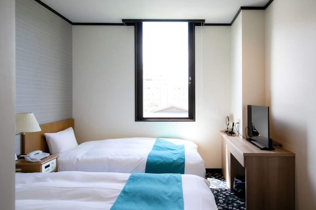 A hotel room with two beds, a desk, a TV, a window, and a lamp.