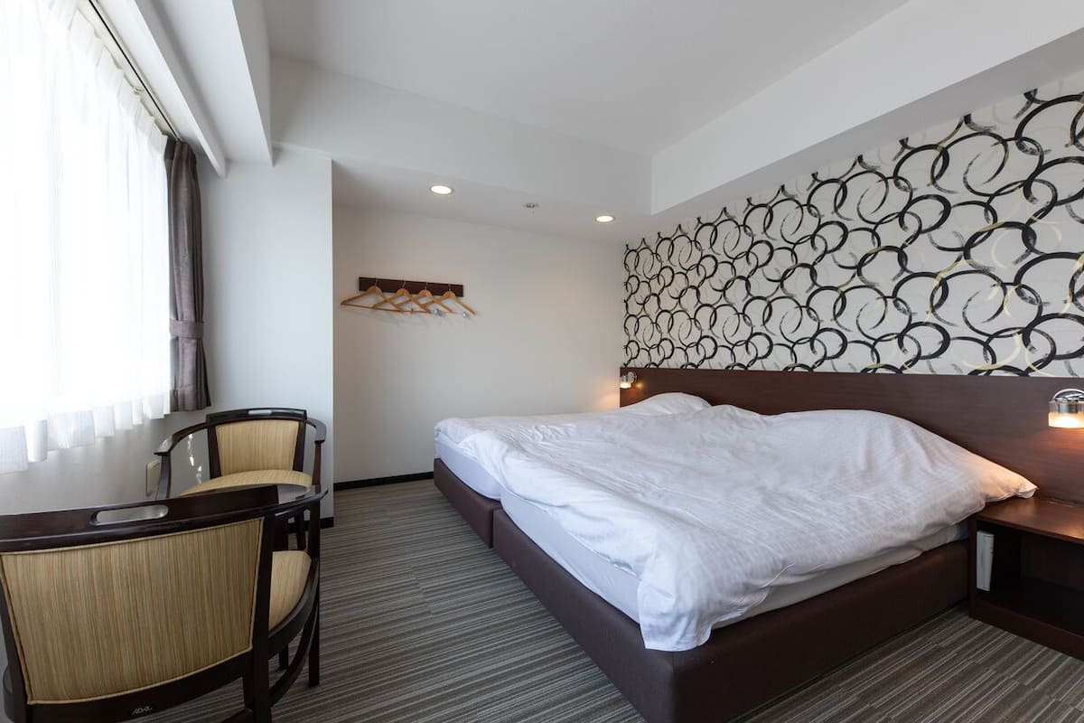 A hotel room with a bed, a desk with chairs, a wall-mounted coat rack, and a patterned wall decoration.