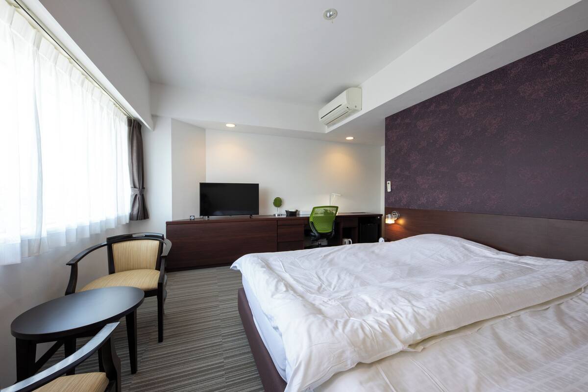 A hotel room with a bed, a desk, a chair, a television, and an air conditioning unit.