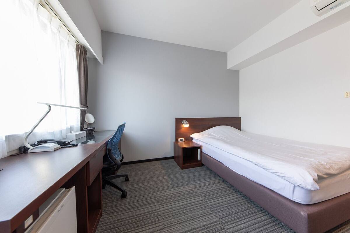 A hotel room with a bed, desk, chair, and a nightstand with a lamp.