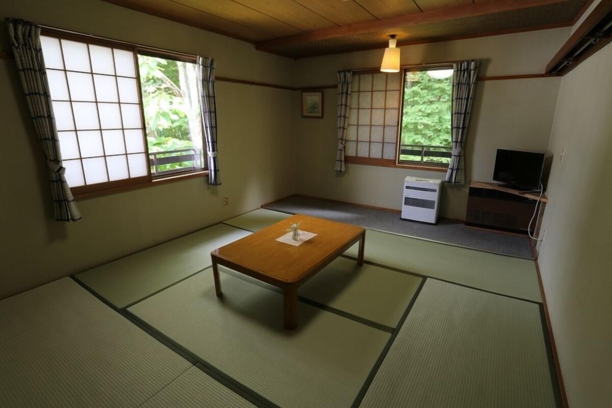 Una habitación tradicional japonesa con suelo de tatami, una mesa de madera baja, un televisor y puertas correderas con cortinas.