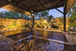 Un área de aguas termales con una pérgola de madera, piso de piedra y grandes rocas.