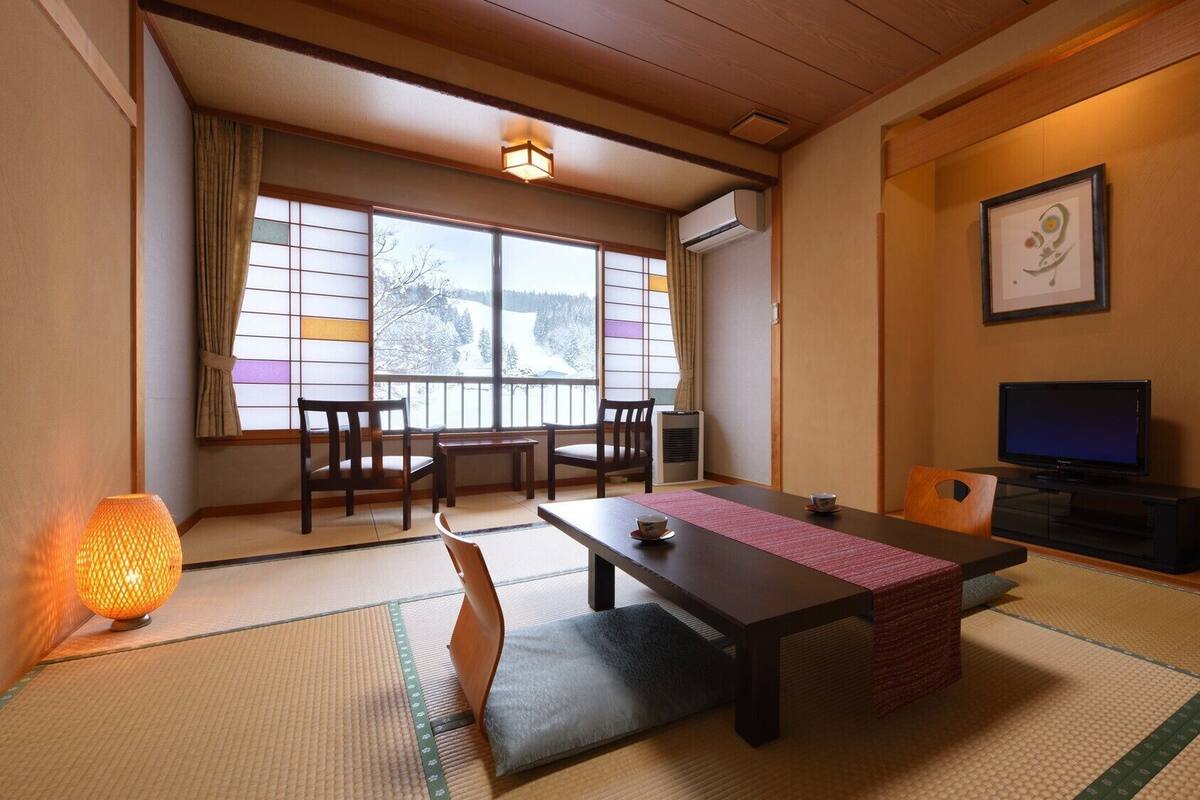 Una habitación japonesa tradicional con suelo de tatami, una mesa baja con cojines, televisión y vista a un paisaje nevado.