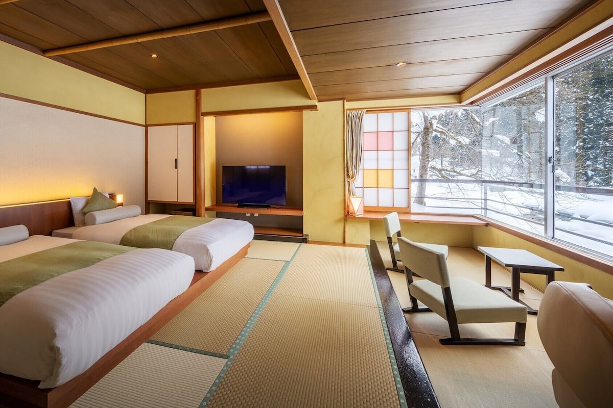 Habitación de hotel con dos camas, televisor, ventana que da a árboles y esteras de tatami.