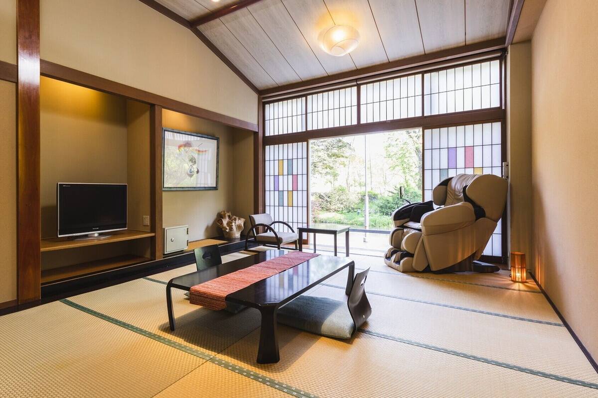 Una habitación japonesa tradicional con suelo de tatami, televisión, sillón de masaje y un ventanal con vistas a la vegetación.