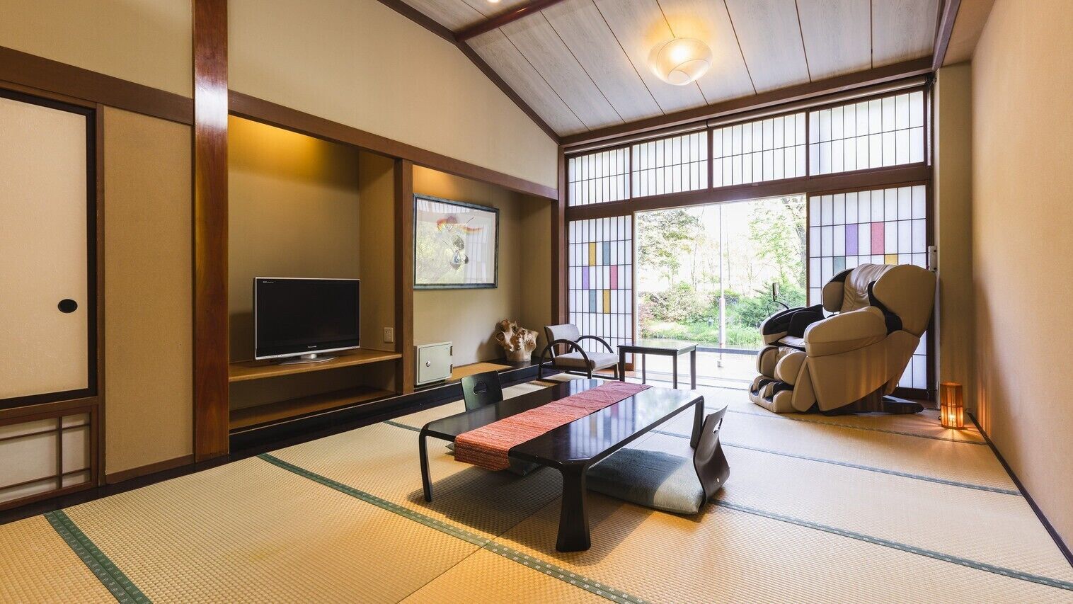 A traditional Japanese room with tatami flooring, a television, a tatami mat table, a sofa, and a large window with a view of greenery.