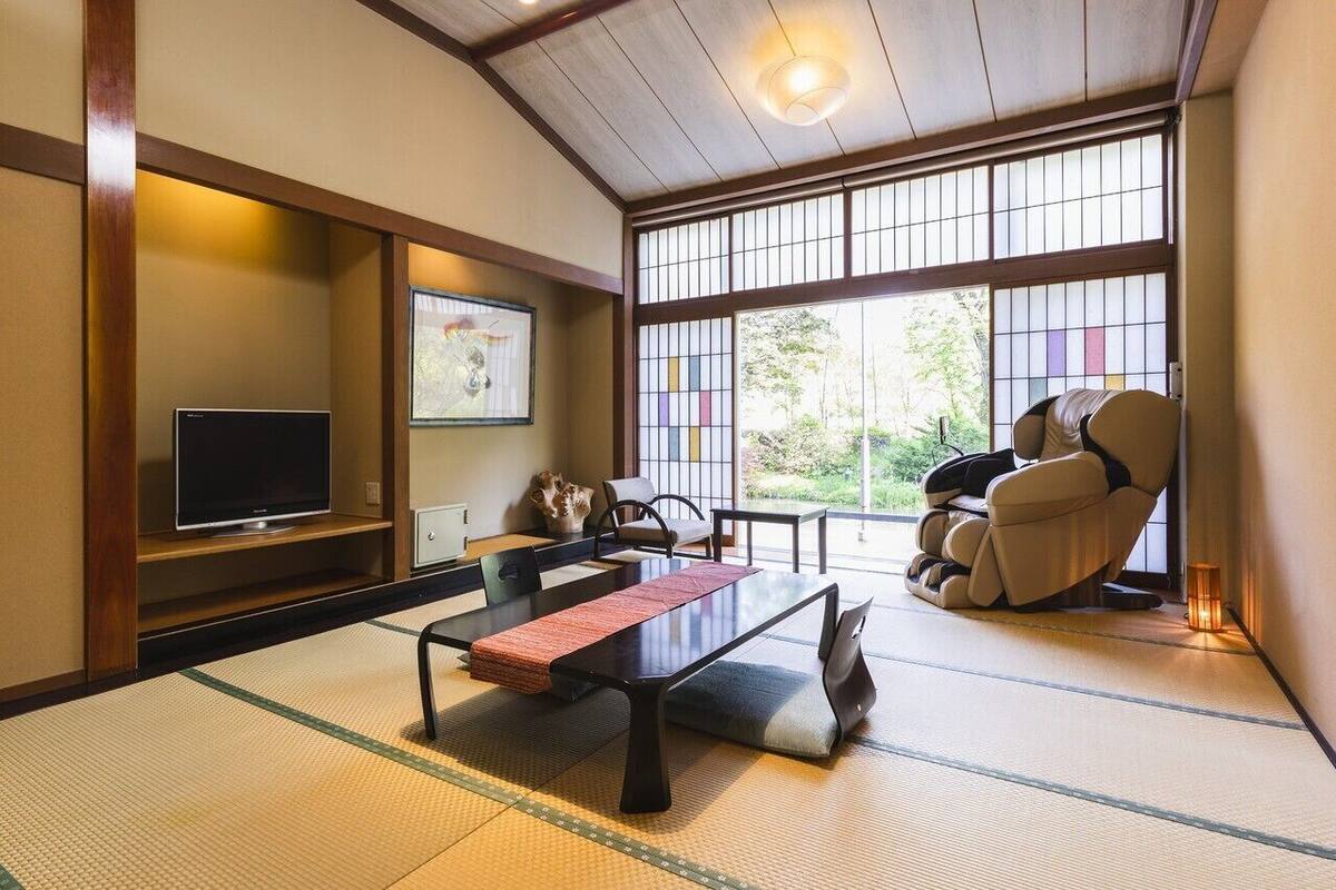 Una habitación japonesa tradicional con suelo de tatami, televisión, una mesa baja de tatami, sofá y un ventanal con vistas a zonas verdes.