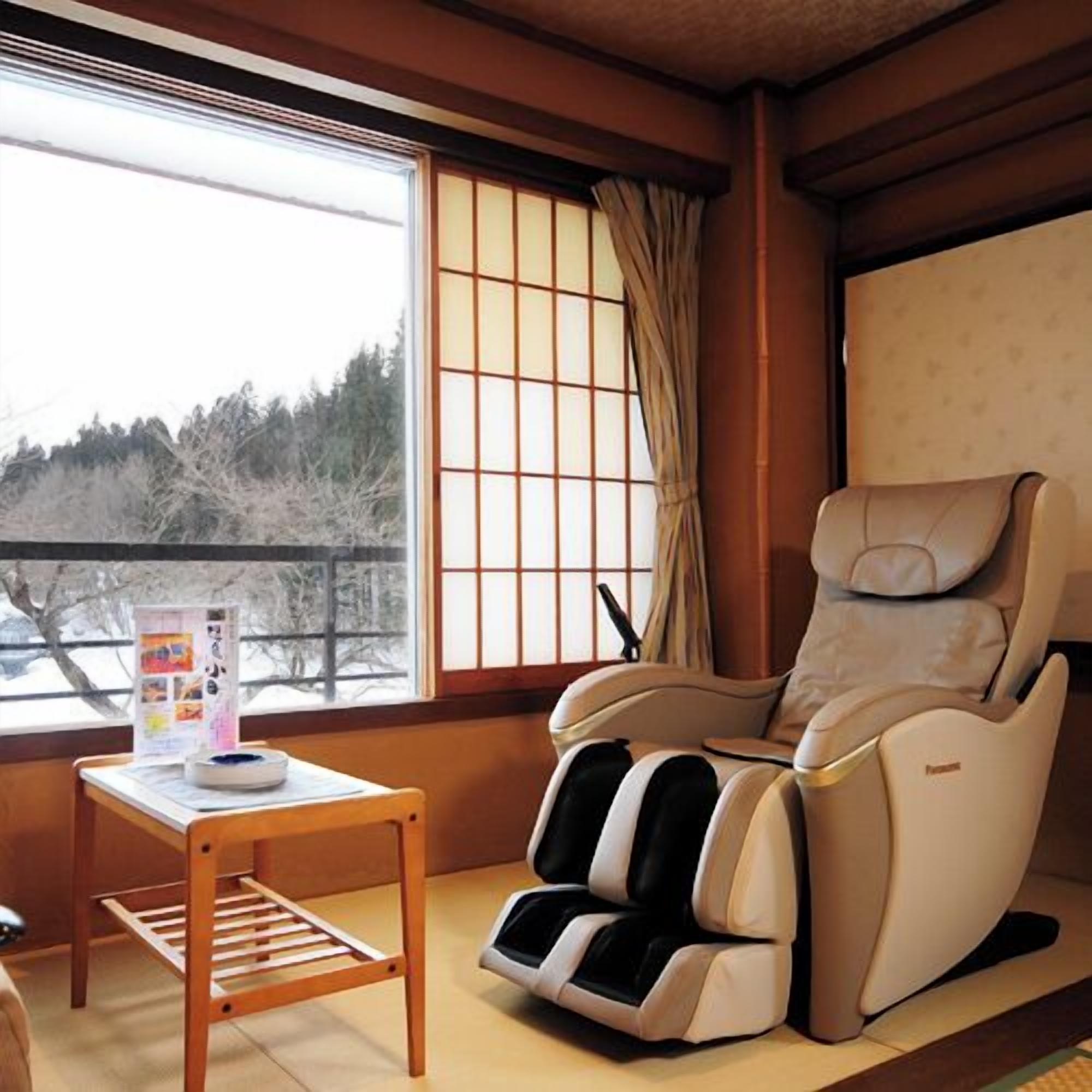 A room with a massage chair, a small table with a magazine, and a window with a view of trees.